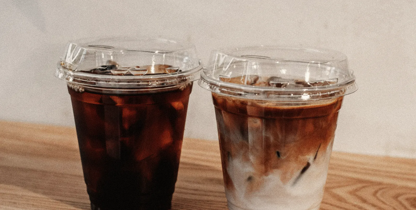 An iced long black and an iced latte side by side in clear cups on a timber ledge