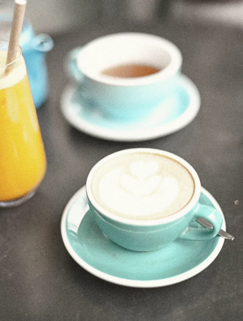 A flat white and a pot of tea in pale blue cups beside an orange juice