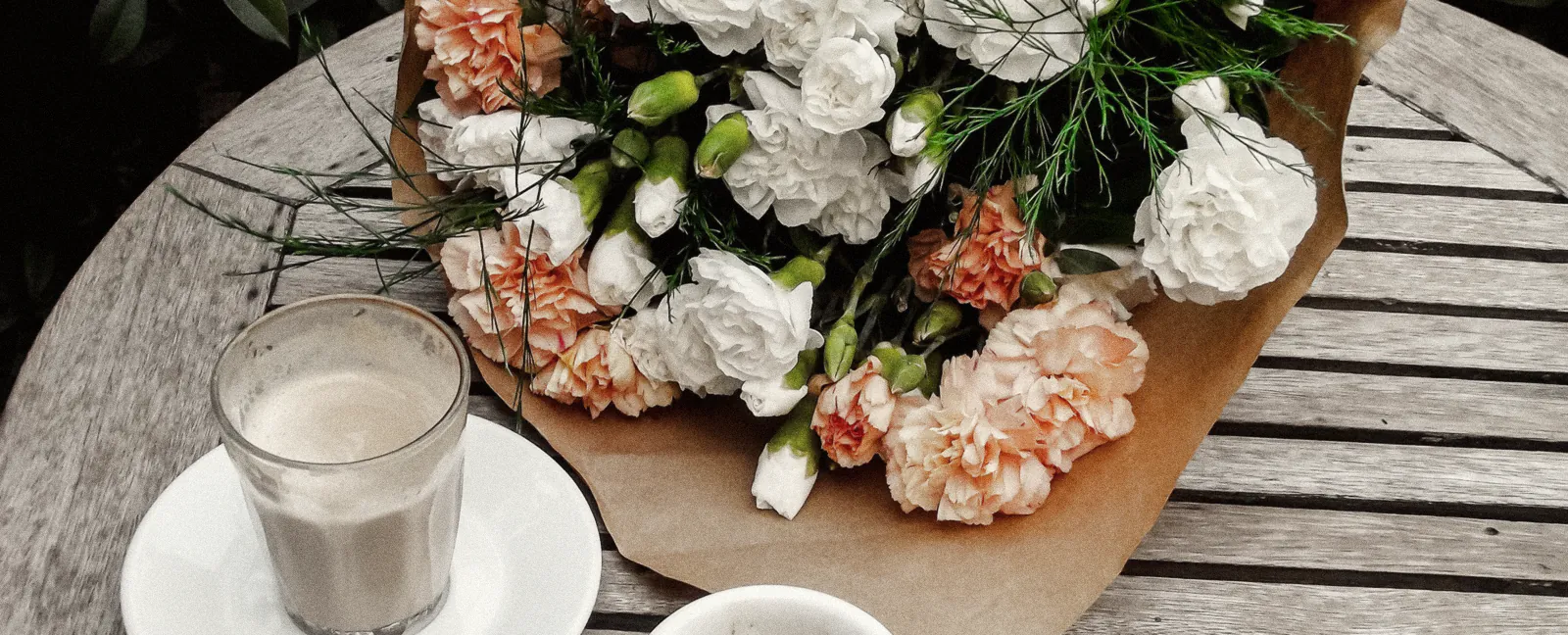 A bunch of peach and white carnations in kraft paper on a slatted courtyard table, beside a glass of latte