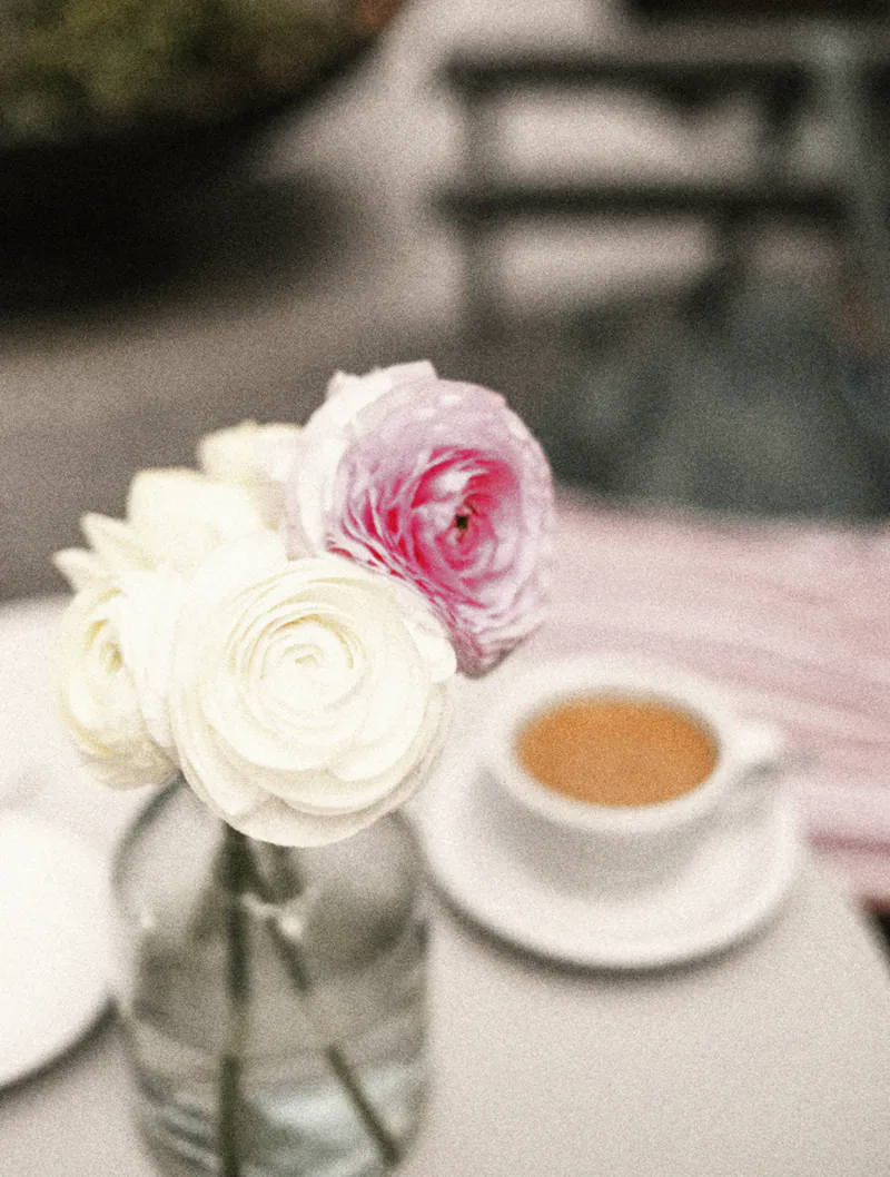 A cup of tea beside white and pink ranunculus on a courtyard table