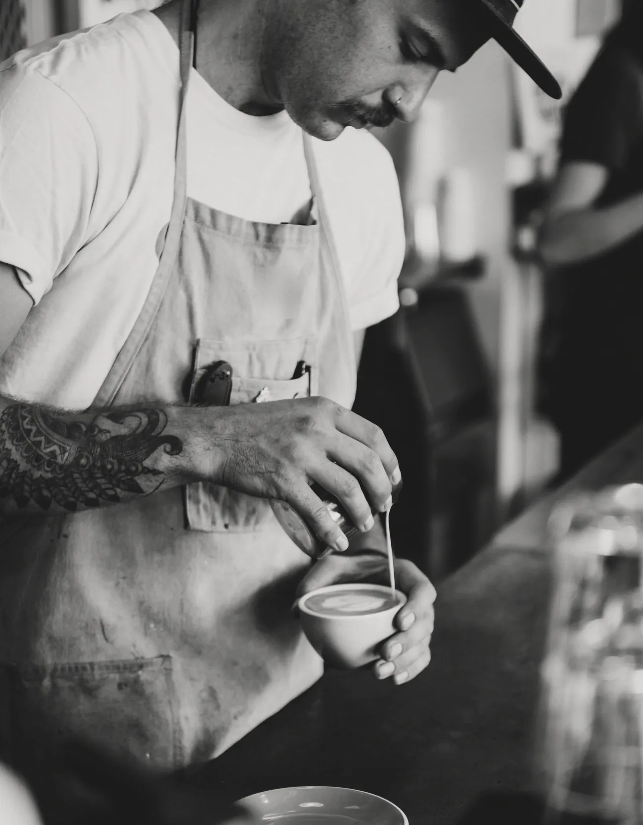 A barista in a cap and apron pours latte art into a cup