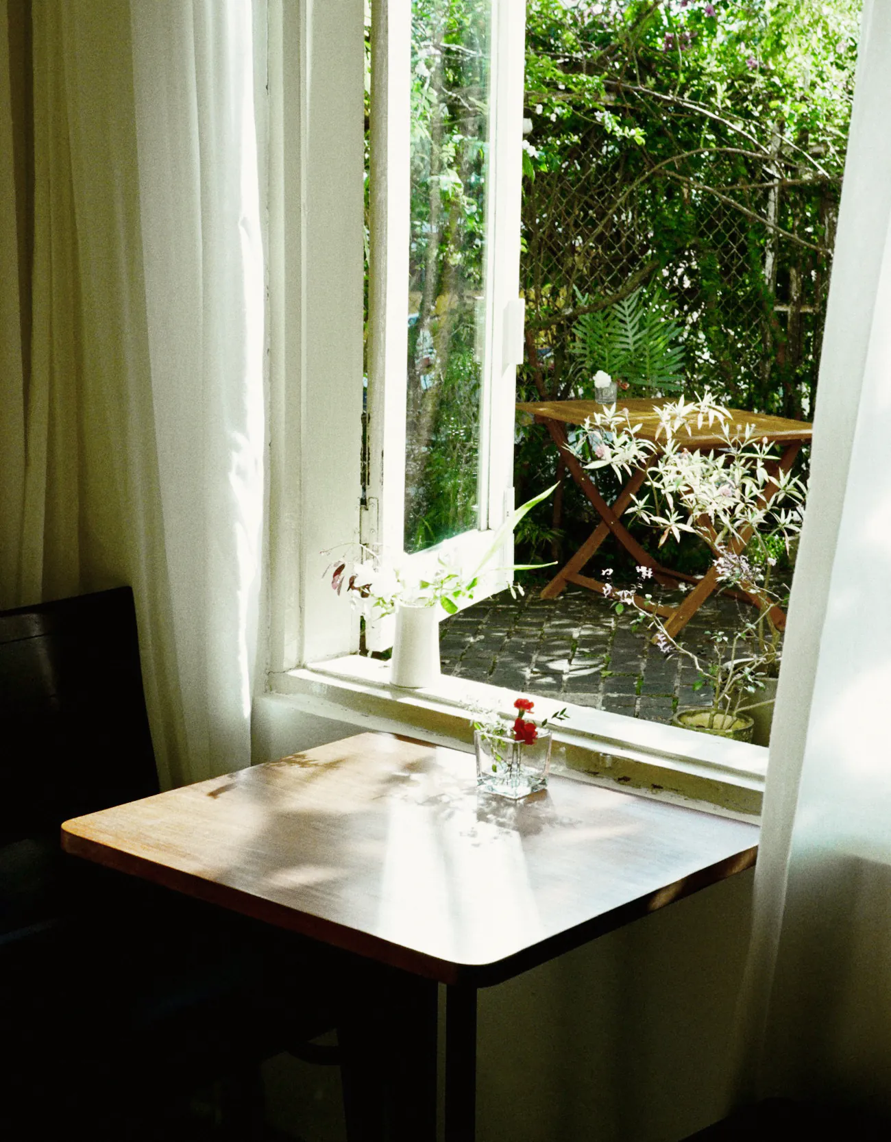 A small table by an open window onto a green courtyard, a bud vase on the sill